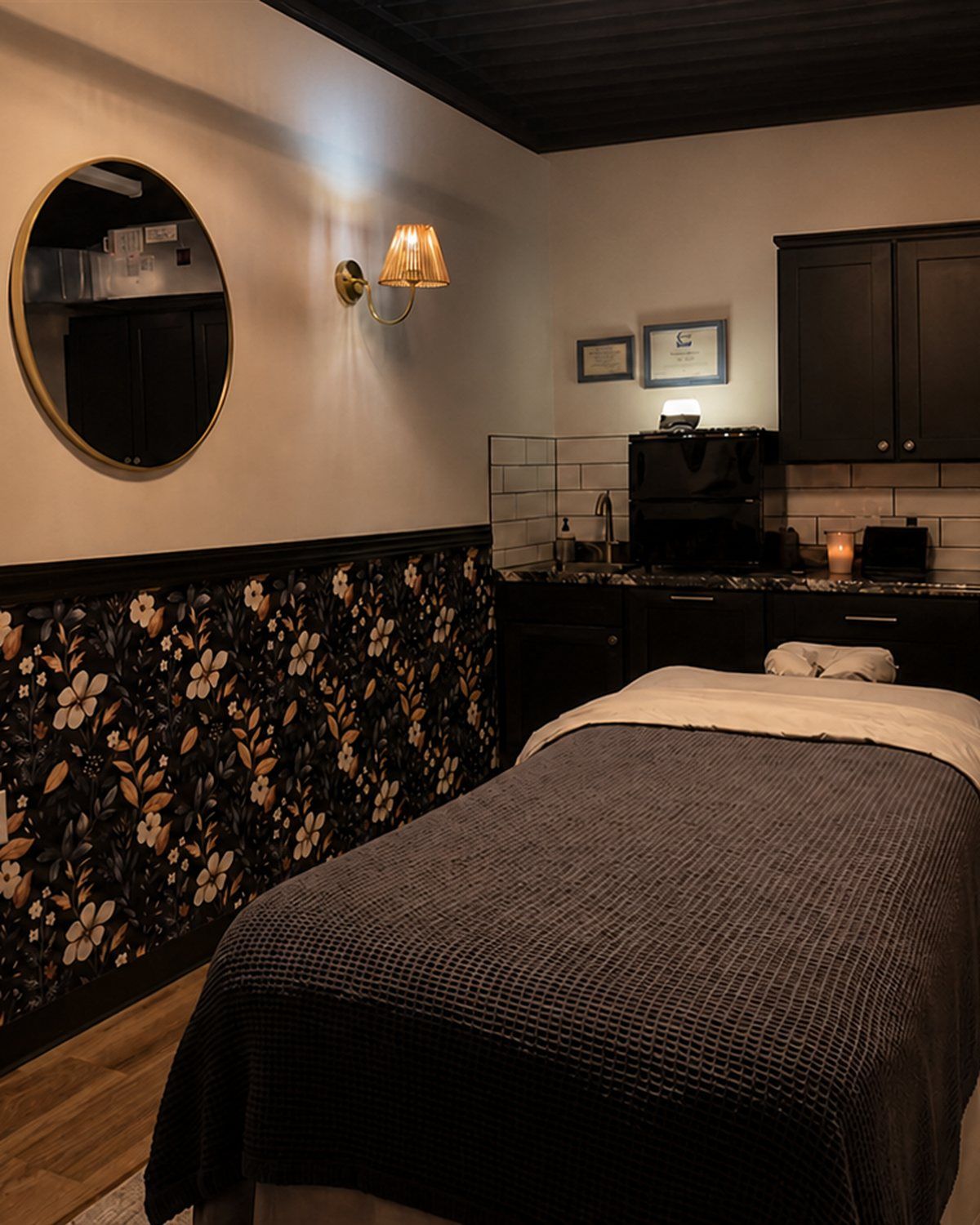 A candlelit Von Rae spa suite with massage table, floral wainscoting, and black cabinetry